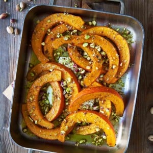 A metal roasting pan filled with roast squash, feta, and pistachios on a wooden table with pistachios scattered around it.