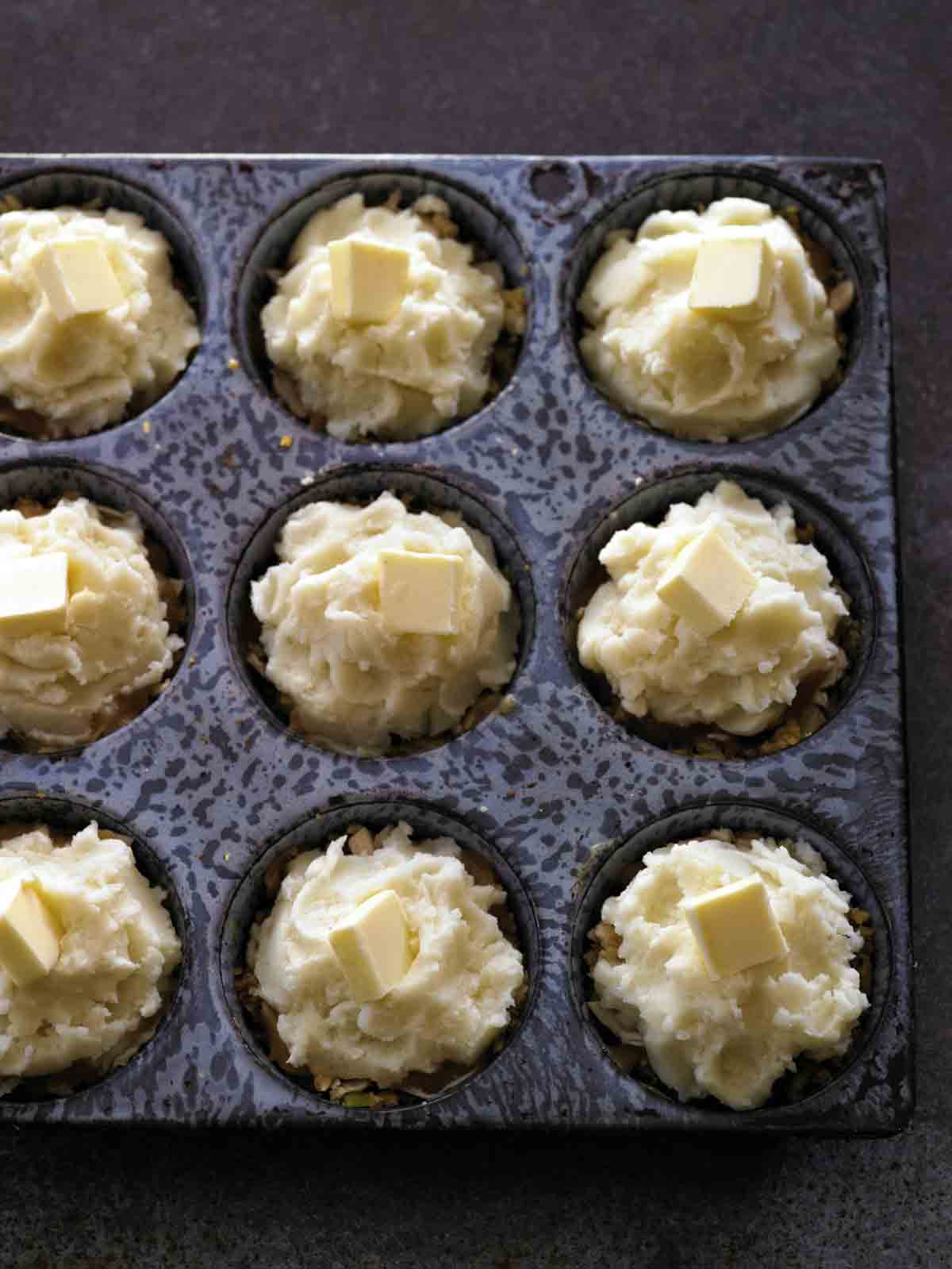 A muffin tin filled with uncooked Thanksgiving leftovers muffins, each topped with a pat of butter.
