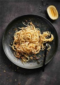 A black plate topped with angel hair pasta with lemon bread crumbs and a fork and a squeezed half lemon rests beside the plate.