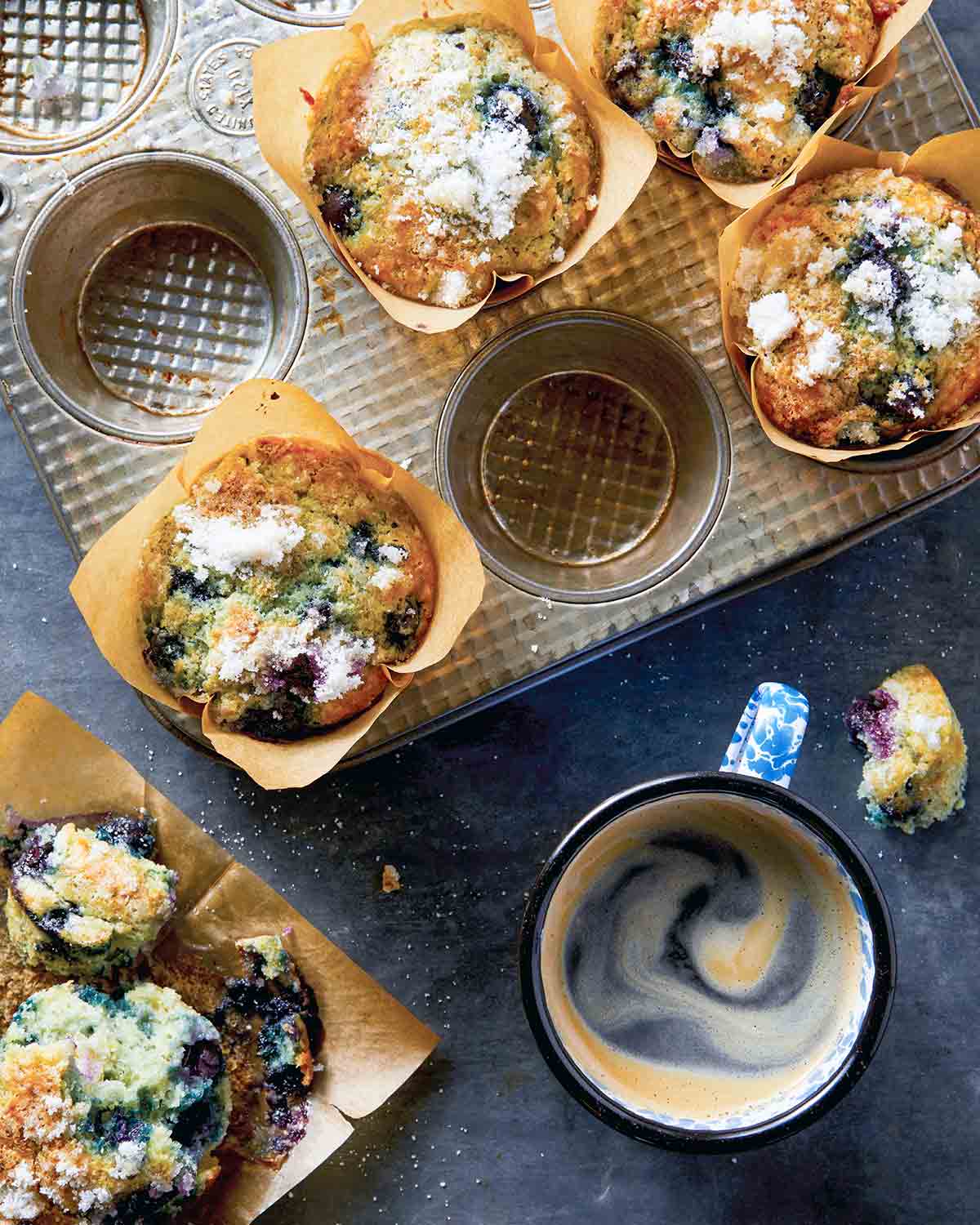 A muffin tray with several blueberry muffins with sugar topping in tulip-style liners, with a halved muffin and a cup of coffee on the side.