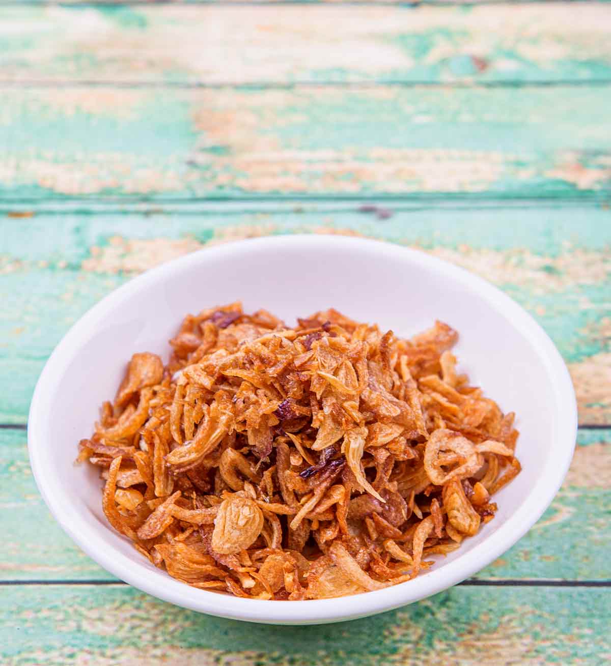 A white bowl filled with crispy fried shallots on a blue wooden surface.