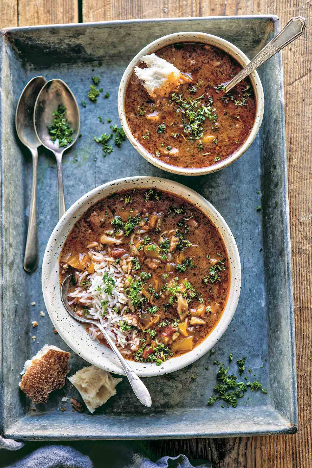 Two bowls of Instant Pot chicken gumbo in a metal tray with spoons, some cubes of bread, and chopped parsley sprinkled on top.