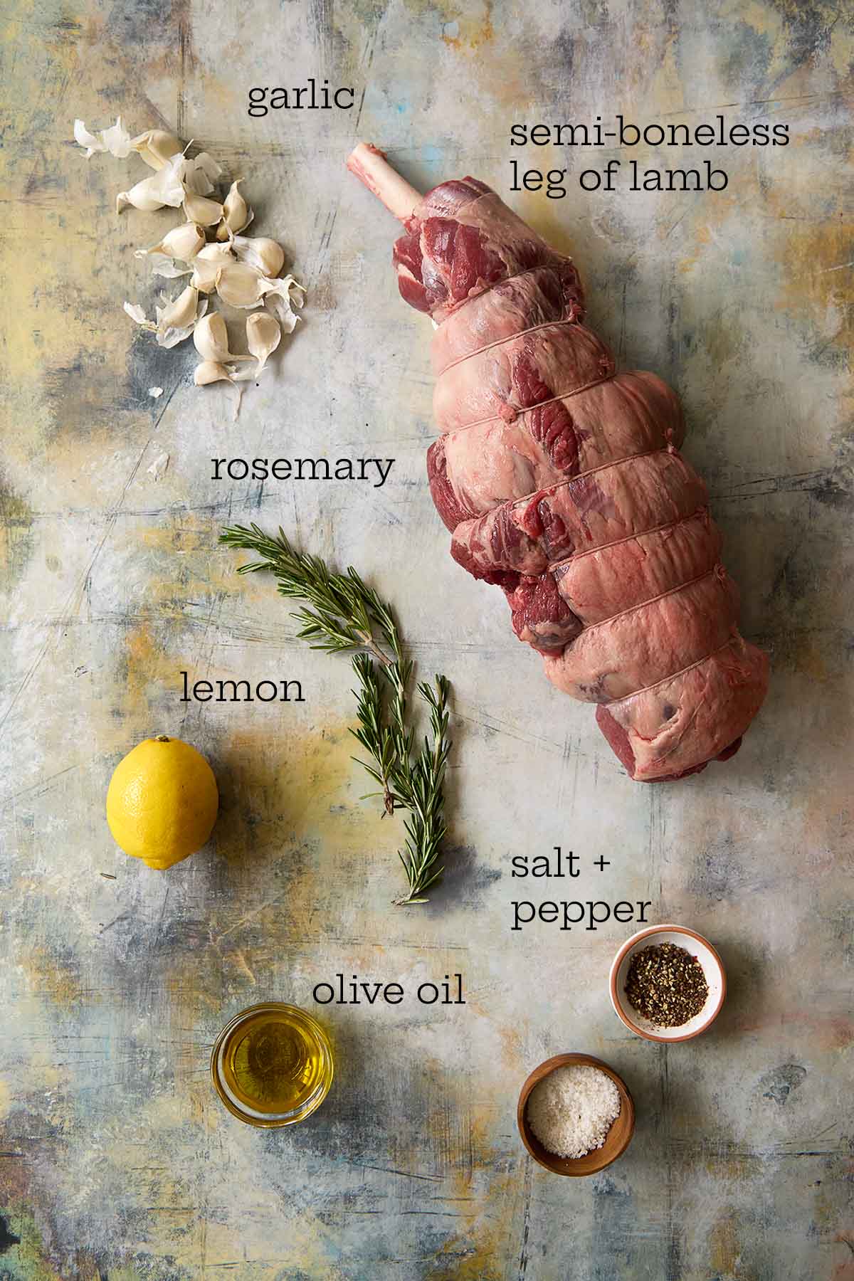 Tied raw leg of lamb on a textured surface with fresh garlic, rosemary, lemon, and olive oil.