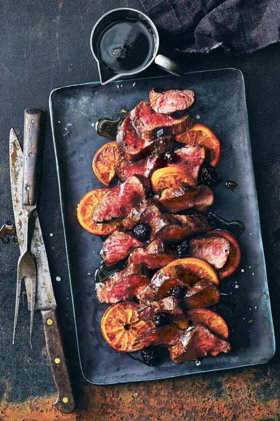 A black tray with sliced roasted venison with balsamic blackberry sauce and sliced oranges.