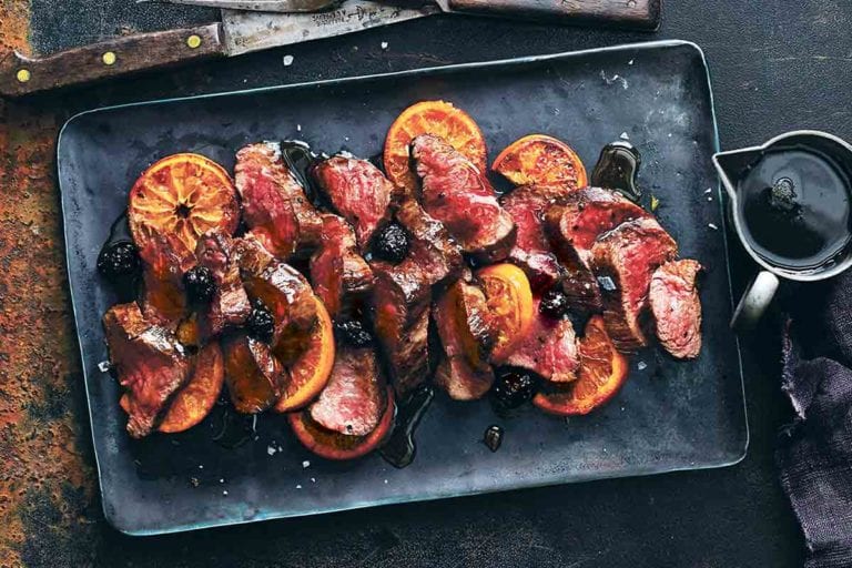 A black tray with sliced roasted venison with balsamic blackberry sauce and sliced oranges.