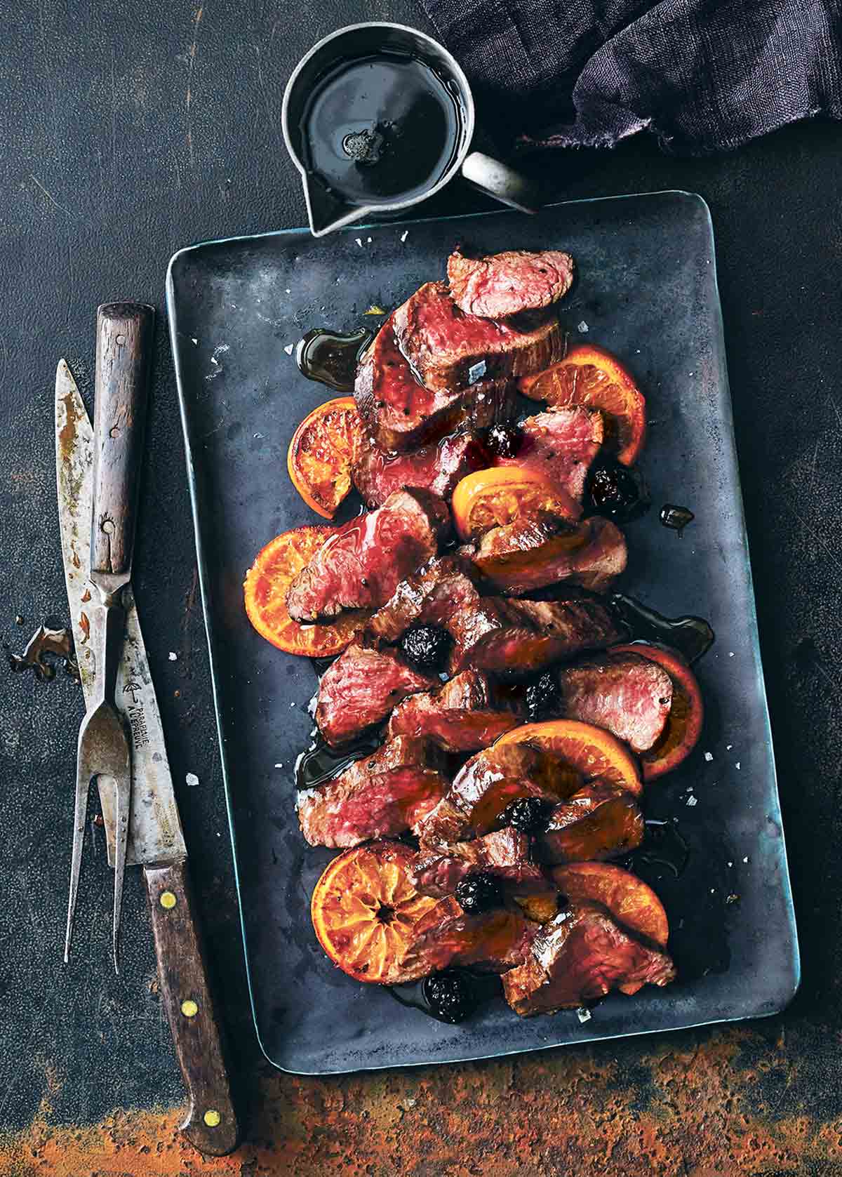 A black tray with sliced roasted venison with balsamic blackberry sauce and sliced oranges.