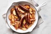 A white enamel cast-iron skillet filled with sautéed sausages with apples on a grey linen cloth.