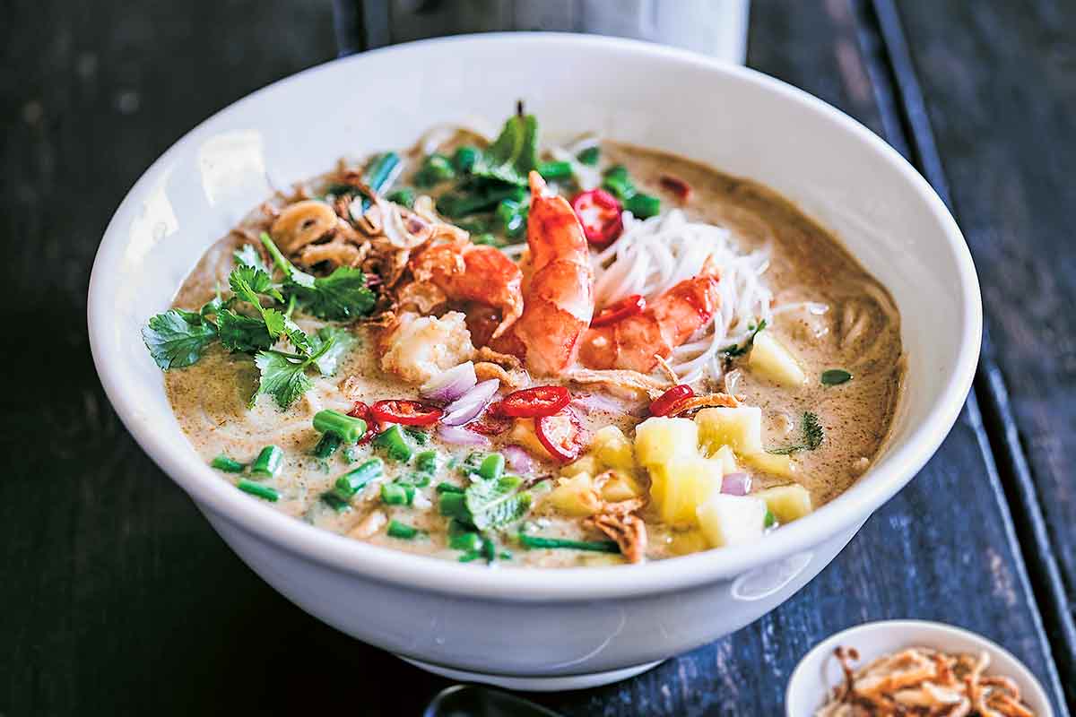 A white bowl of shrimp laksa soup with a small bowl of crispy fried shallots, a spoon, and a container of chopsticks on the side.