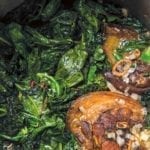 A large stock pot filled with Southern greens with ham hocks and chopped onions.