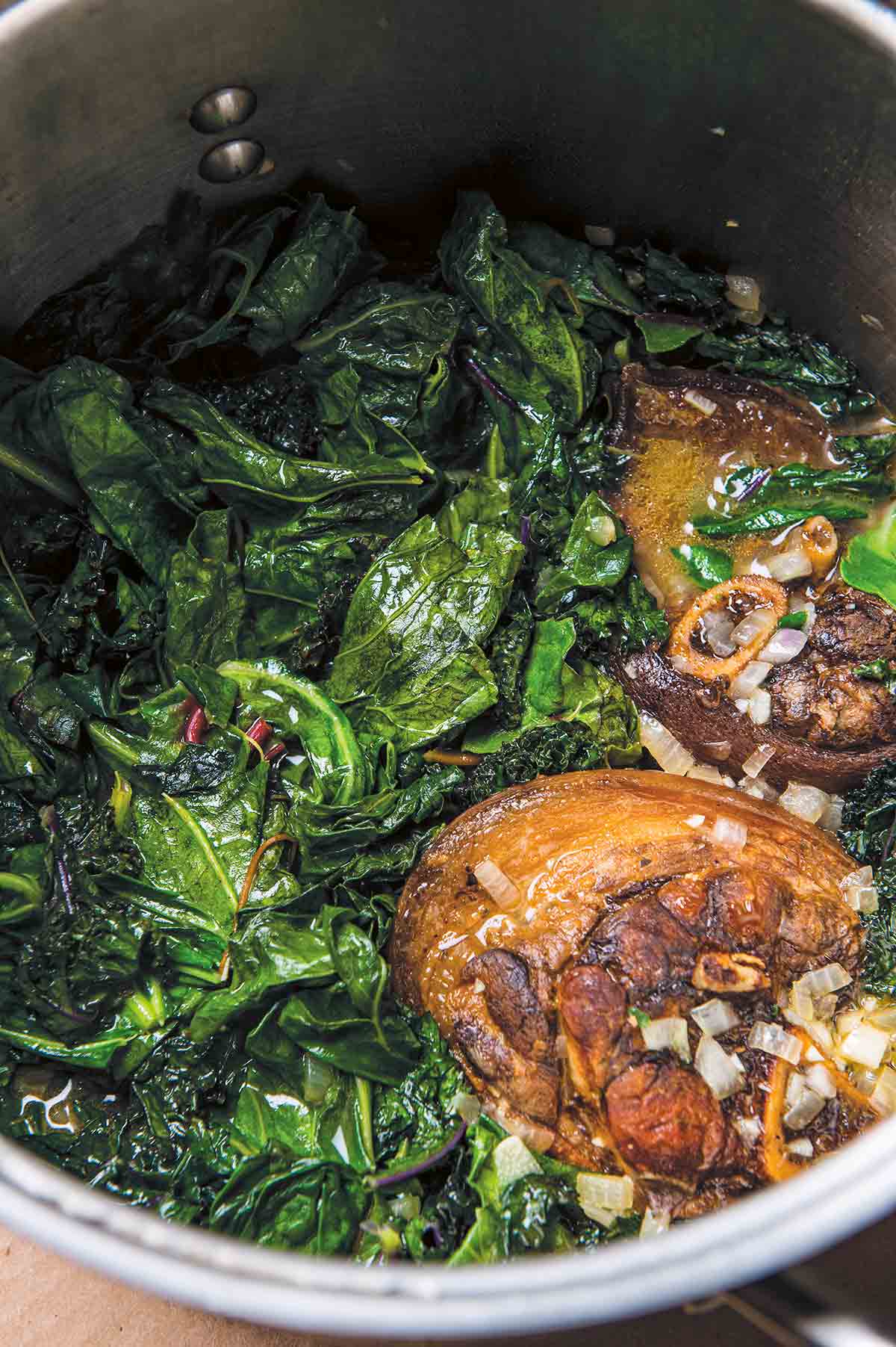 A large stock pot filled with Southern greens with ham hocks and chopped onions.