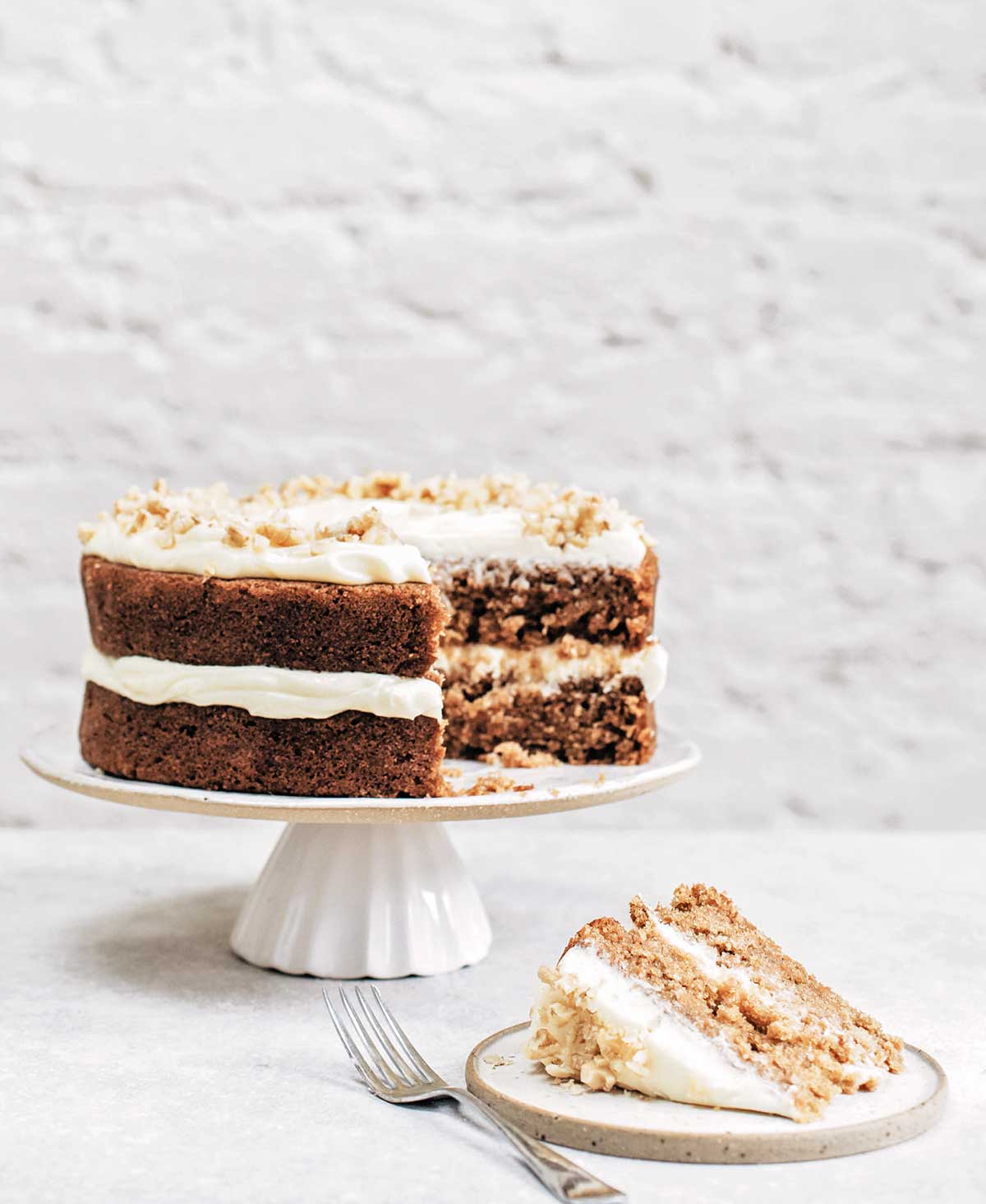 A whole two layer spiced carrot cake on a white cake stand with one slice cut from it on a plate with a fork beside it.