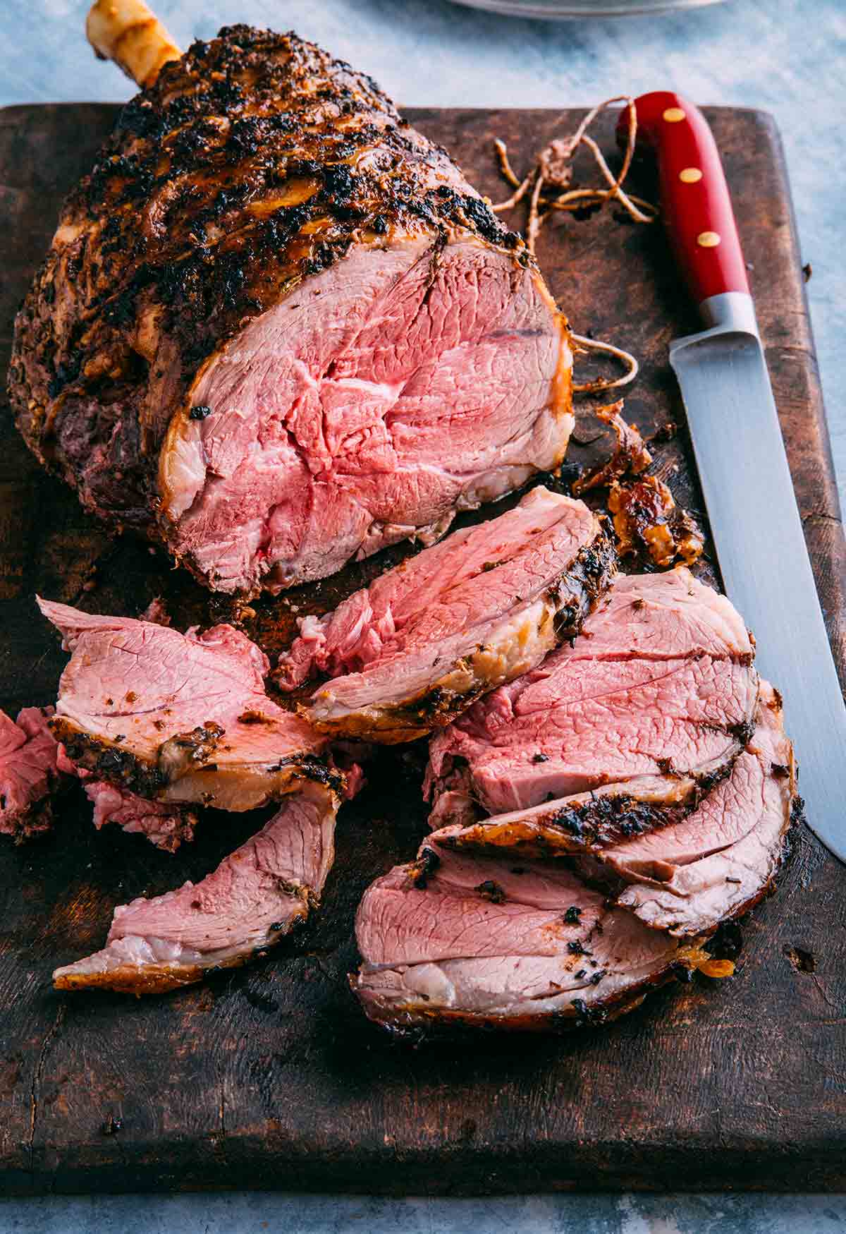 A partially sliced semi-boneless Tuscan style roasted leg of lamb on a wooden cutting board with a knife resting beside the lamb.