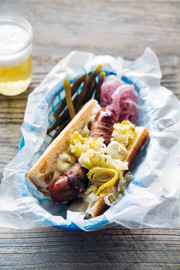 A bacon-wrapped hot dog topped with sauerkraut in a blue, wax paper-lined basket with a glass of beer beside it.