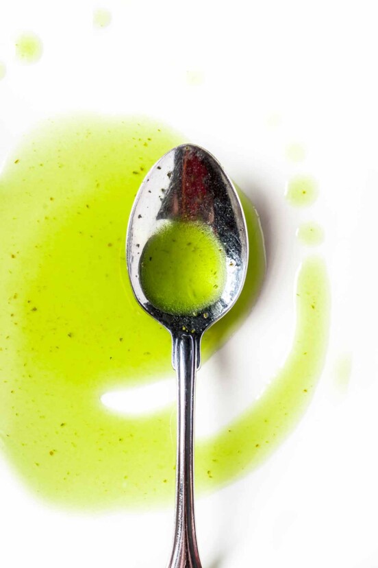A silver spoon resting in a drizzle of basil oil on a white surface.