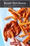 A hand dipping fries into a splatter of bomb hot sauce with a pile of fries scattered beside the sauce.