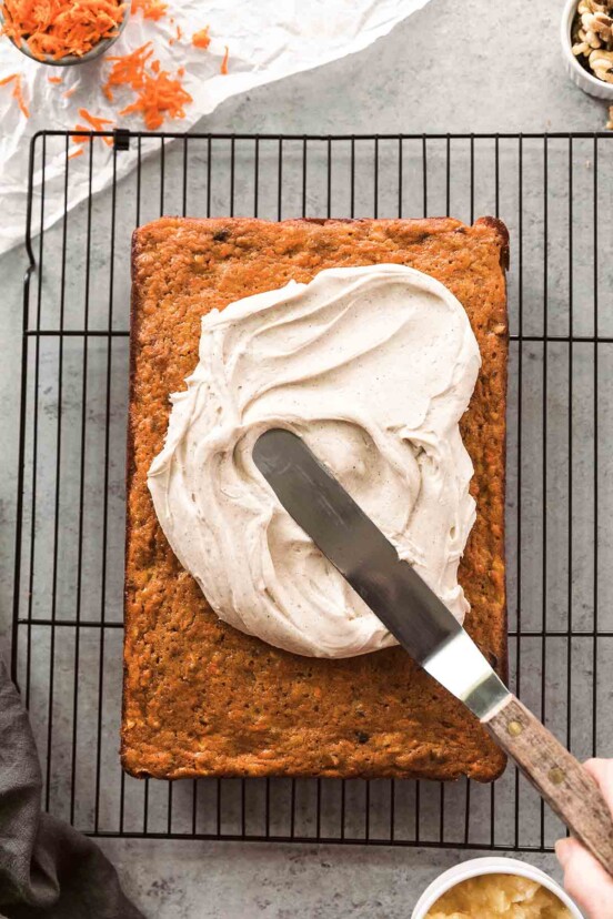 A whole carrot sheet cake on a wire rack with chai cream cheese frosting being spread over the top.