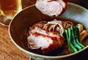 A bowl of duck soba with scallions on a wooden surface with a piece of duck being lifted out with chopsticks.