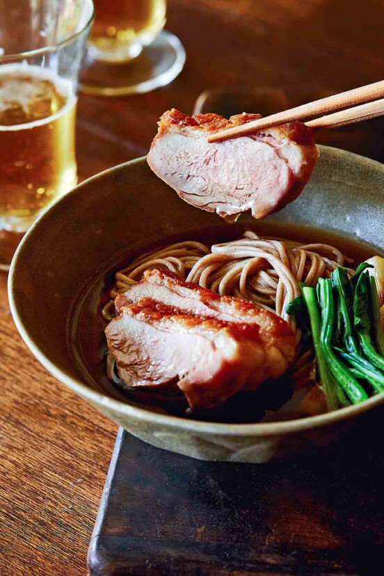 A bowl of duck soba with scallions on a wooden surface with a piece of duck being lifted out with chopsticks.