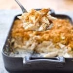 A dark grey rectangular baking dish half filled with four cheese macaroni and cheese and a metal spoon lifting a scoop out.