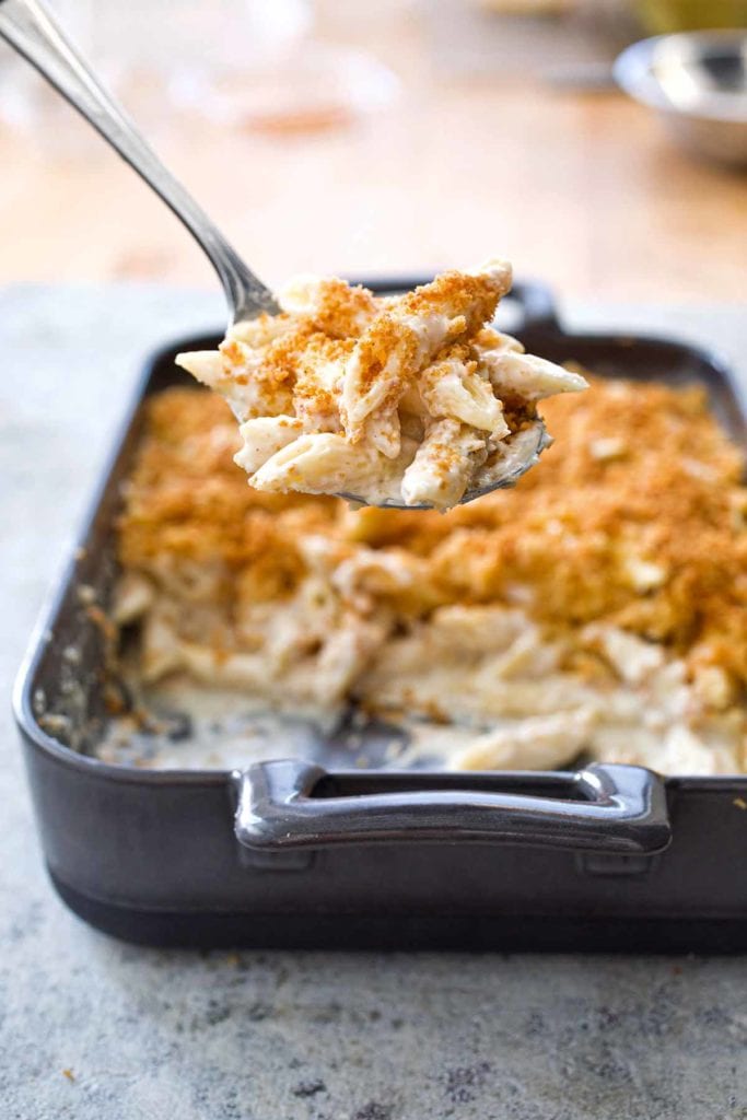 A dark grey rectangular baking dish half filled with four cheese macaroni and cheese and a metal spoon lifting a scoop out.