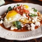 A white plate topped with huevos rancheros - crispy tortilla, fried eggs, fresh salsa, and Cotija cheese.