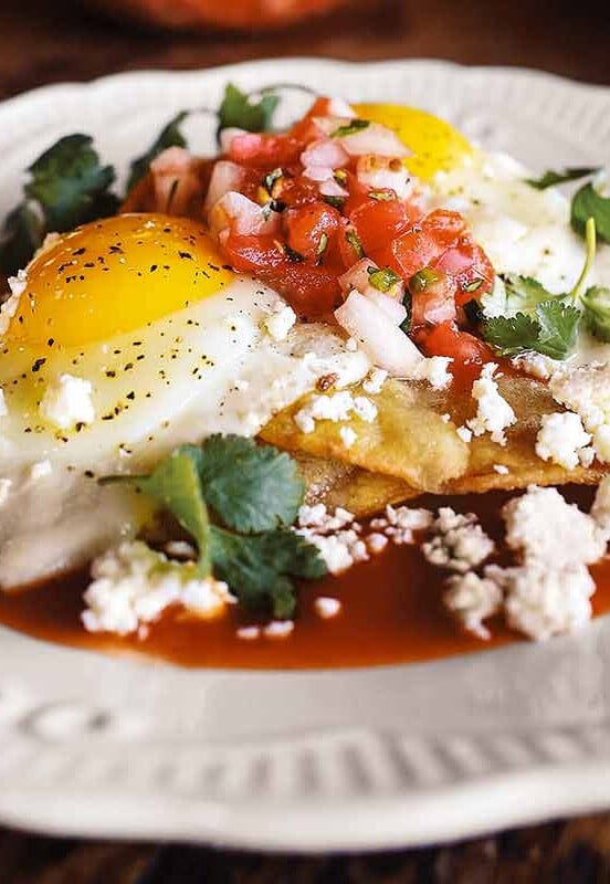 A white plate topped with huevos rancheros - crispy tortilla, fried eggs, fresh salsa, and Cotija cheese.