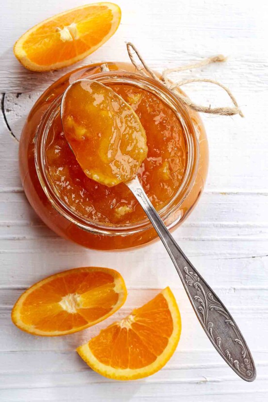 A jar of orange marmalade with a spoon resting on top and a few orange wedges beside it.