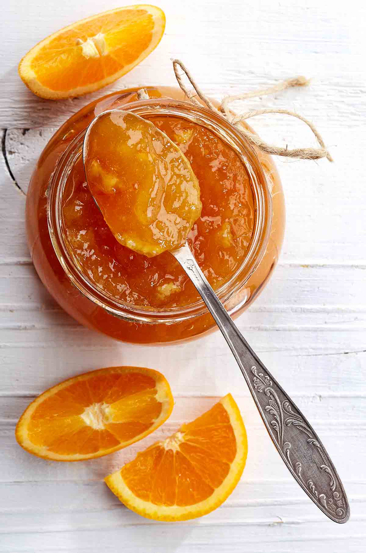 A jar of orange marmalade with a spoon resting on top and a few orange wedges beside it.