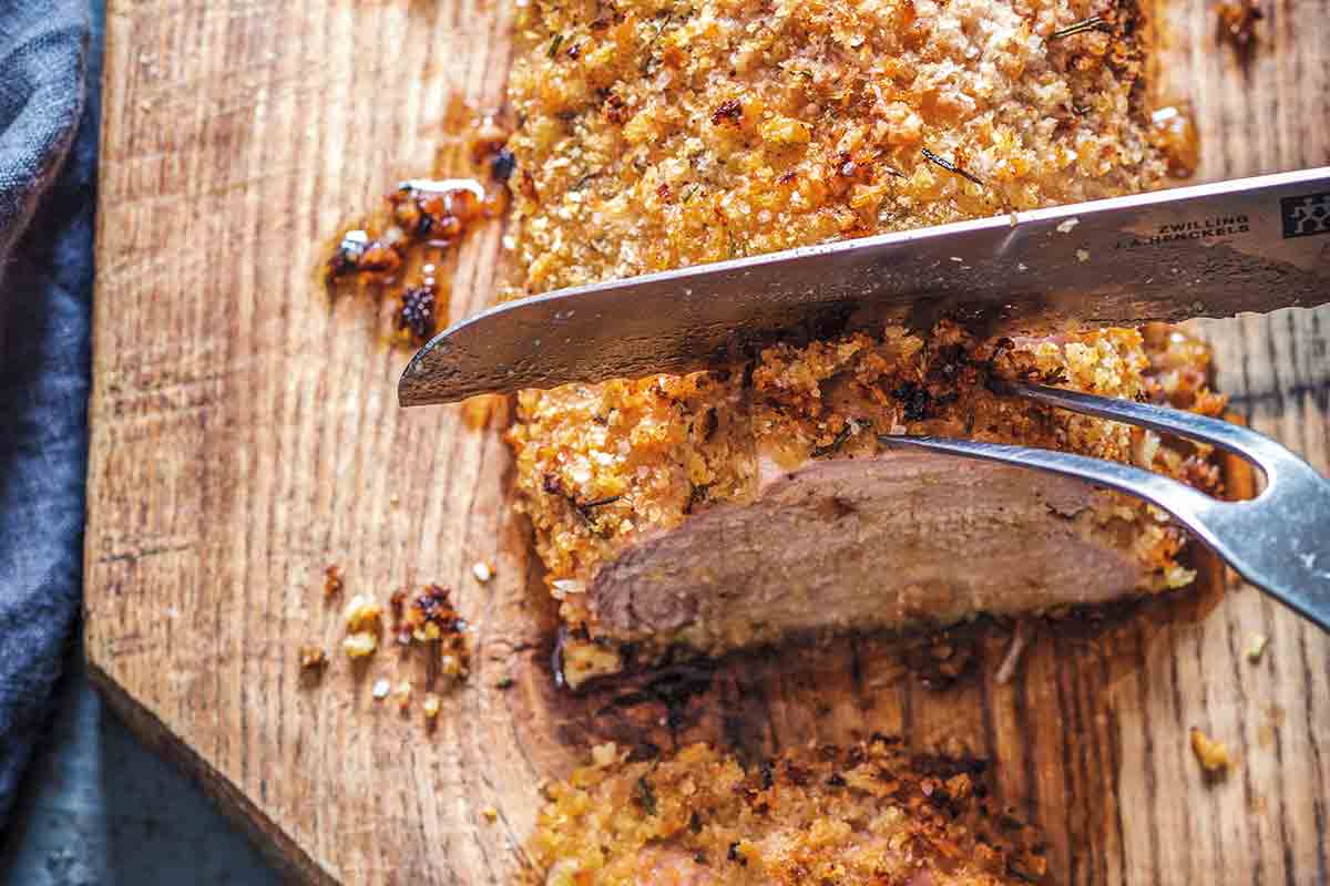 A panko crusted pork tenderloin with rosemary being sliced on a wooden cutting board.