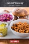 A bowl of oven roasted pulled turkey breast, a bowl of cabbage slaw, a jar of pickles, and several whole wheat buns on a white tray.