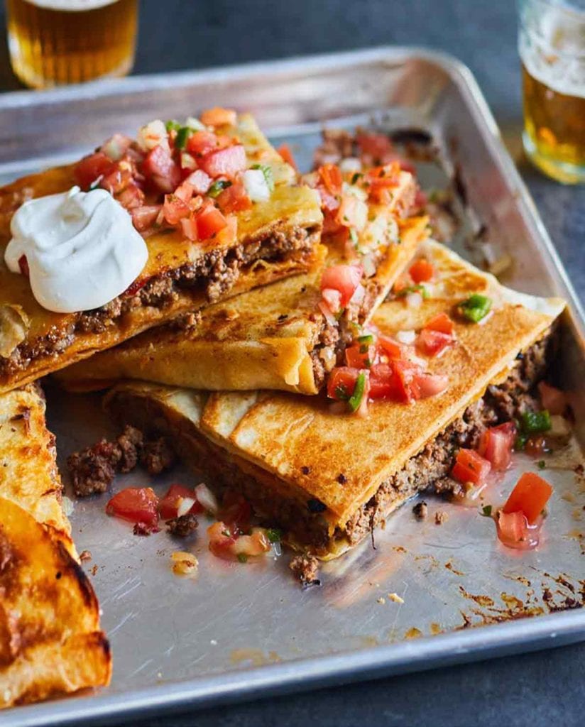 A cut sheet pan taco bake in a rimmed sheet pan topped with pico de gallo and sour cream.