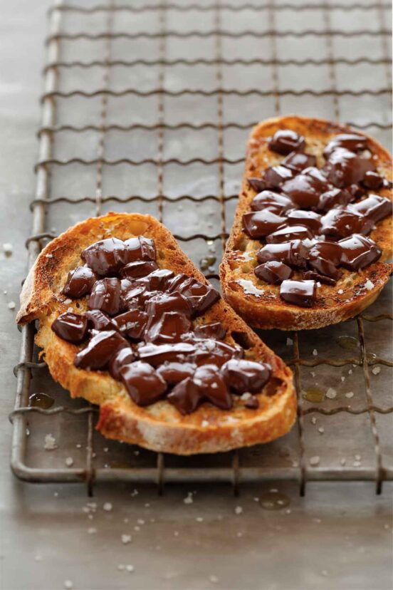 Two golden brown slices of sourdough toast topped with melting chunks of chocolate and a sprinkling of chunky sea salt on a rack.