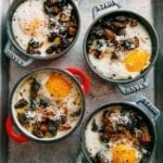 Four individual cocottes filled with baked eggs with polenta and mushrooms on a rimmed baking sheet.