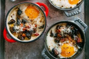 Four individual cocottes filled with baked eggs with polenta and mushrooms on a rimmed baking sheet.