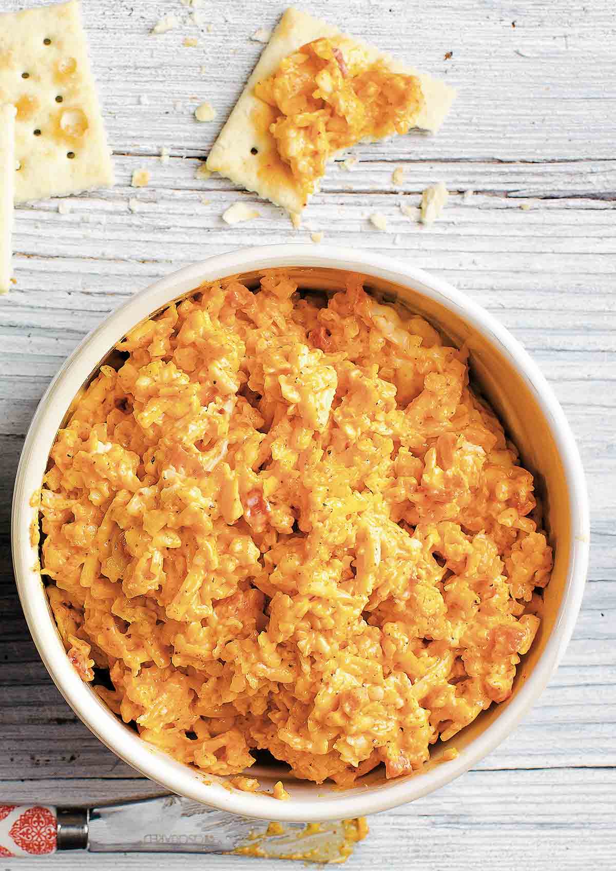 A white bowl filled with pimento cheese and some saltine crackers topped with the cheese next to the bowl