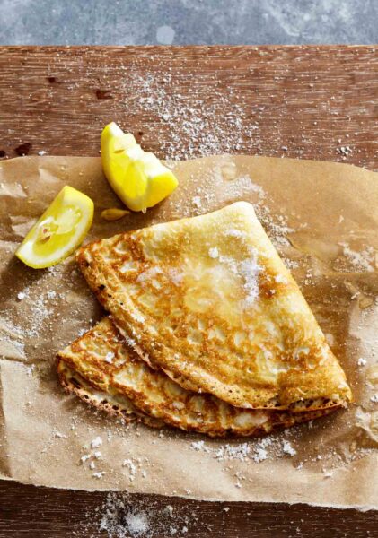 A single basic crepe, folded into quarters on a sheet of parchment with two lemon wedges beside it.