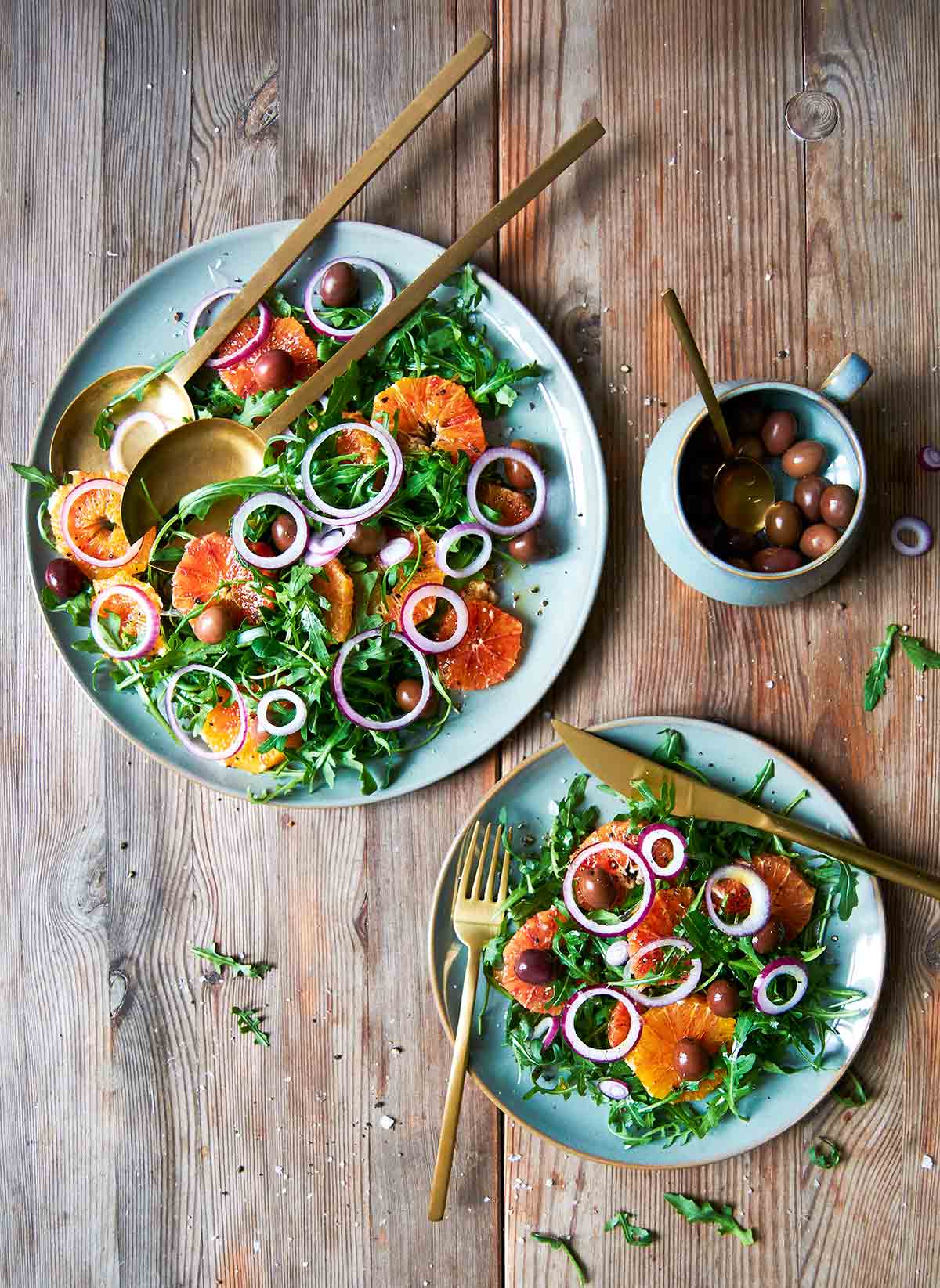 A large platter and smaller plate topped with blood orange and arugula salad with a dish of black olives on the side.