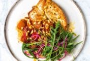 A white plate topped with a piece of brown butter flattened chicken, tomato sauce, arugula and red onion salad, and pine nuts.