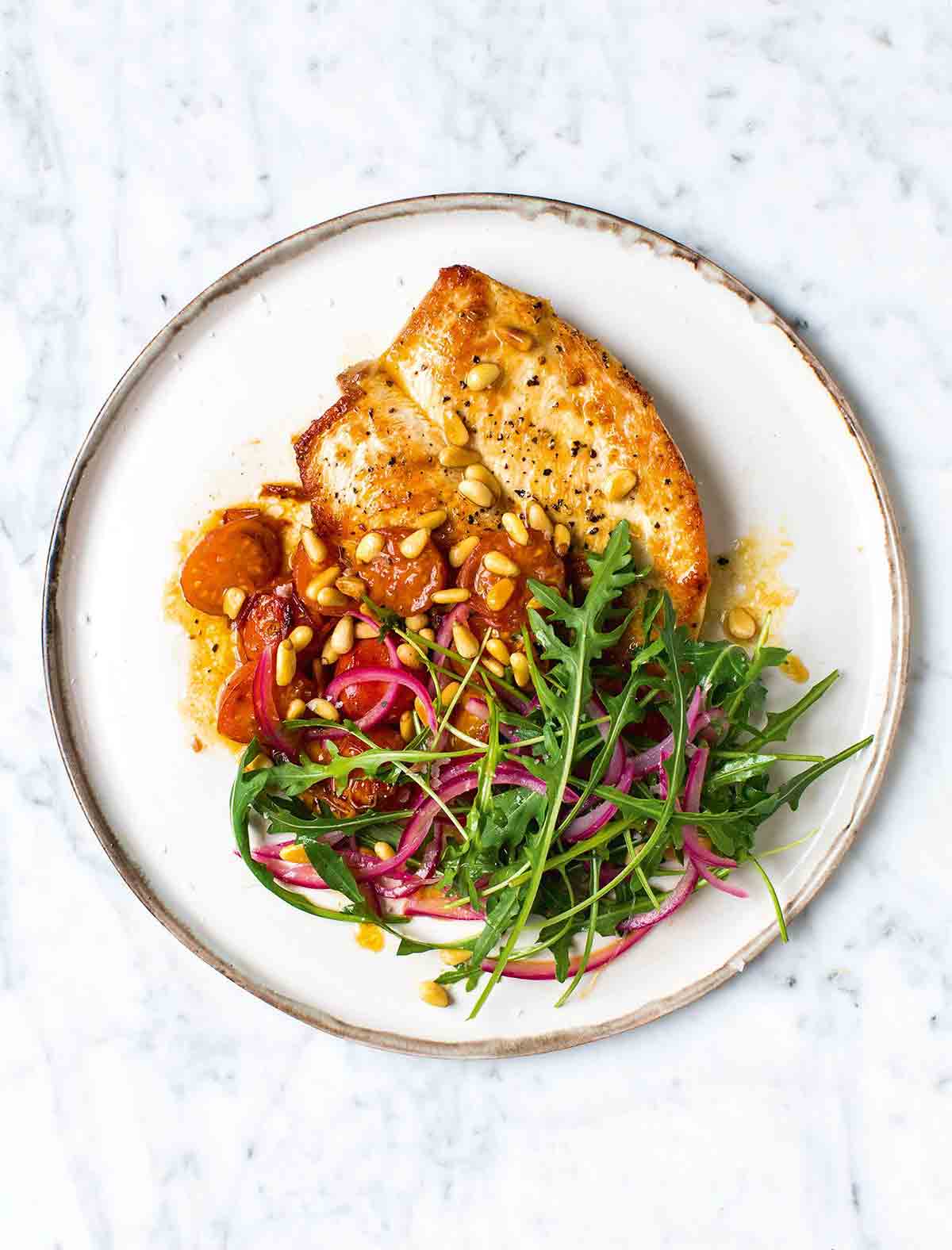 A white plate topped with a piece of brown butter flattened chicken, tomato sauce, arugula and red onion salad, and pine nuts.