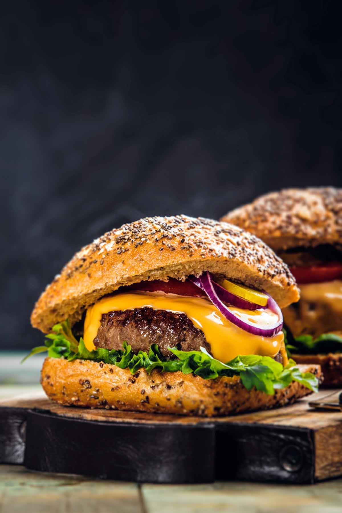 Two cheeseburgers with red onion, lettuce, and pickles on sesame buns on a wooden serving board.