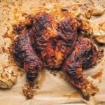 A flat roasted chicken with smoked paprika on a sheet of parchment paper with a halved head of garlic on either side.