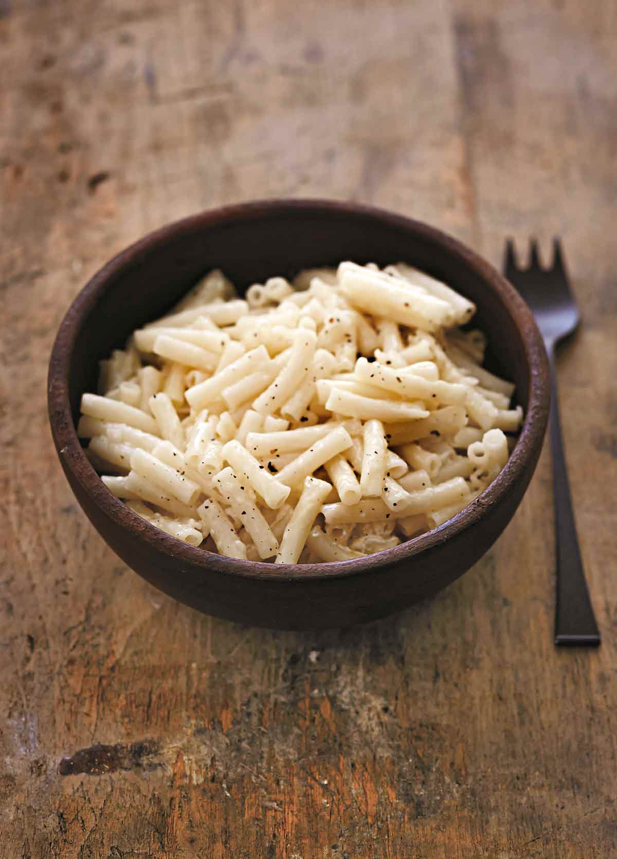 A brown bowl filled with gluten-free macaroni and cheese sprinkled with pepper on a wooden table with a fork resting beside the bowl.