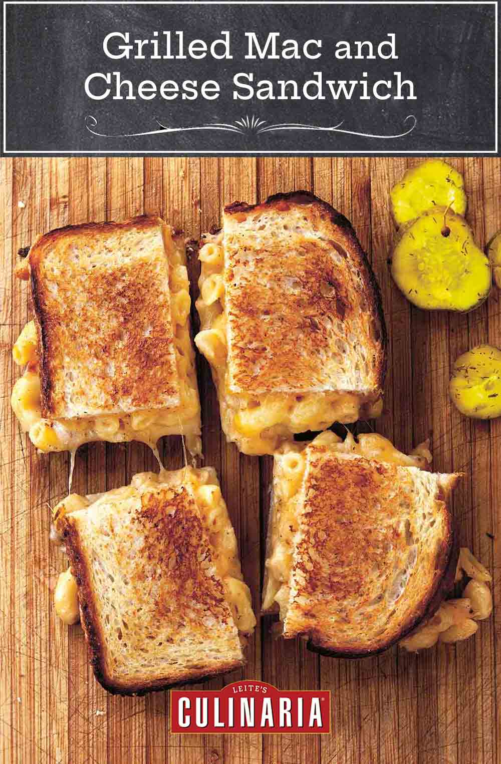 A grilled mac and cheese sandwich cut into four squares on a wooden board with pickle slices beside it.
