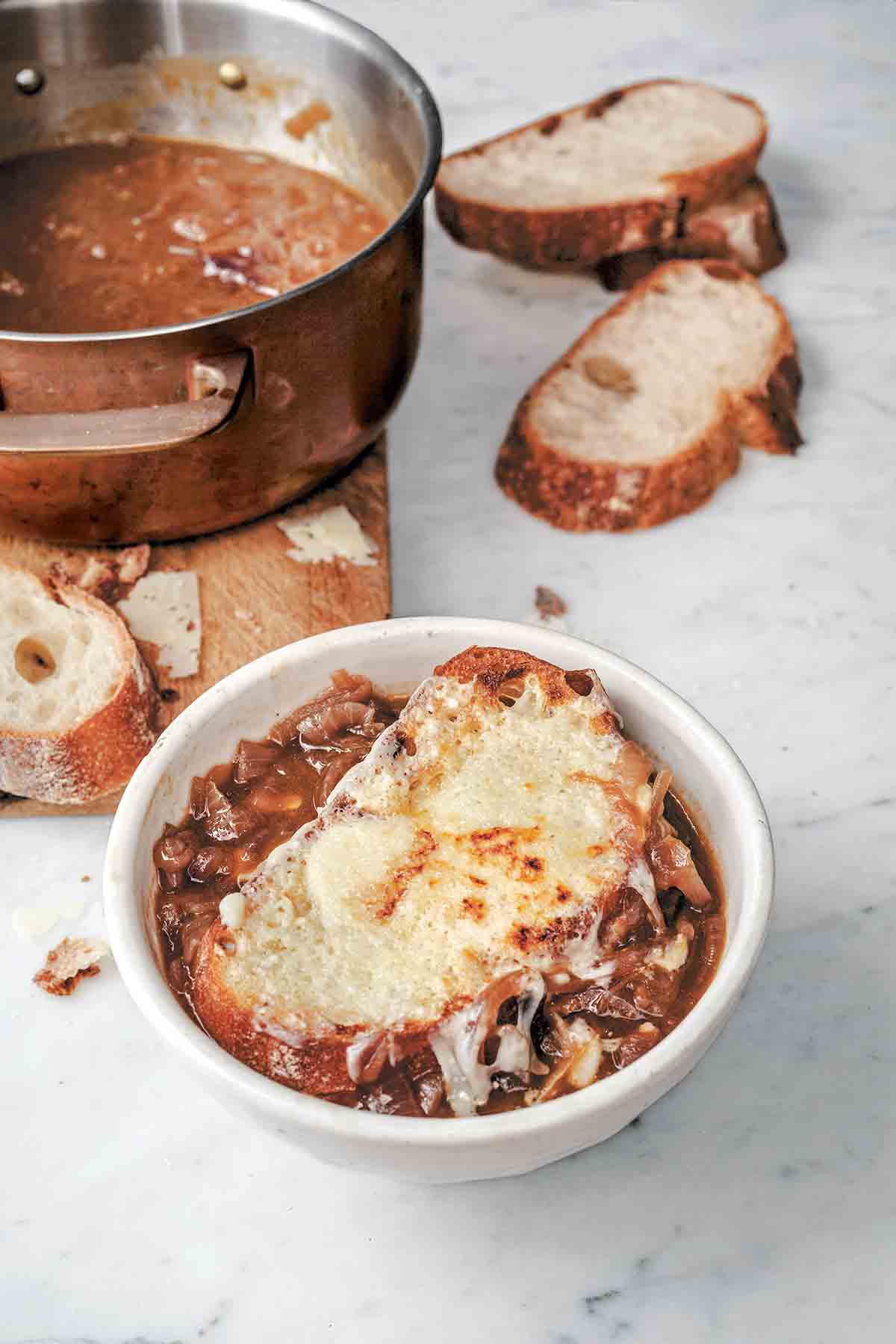 A white bowl of Italian onion soup topped with cheese toast with a pot of soup and several bread slices in the background.