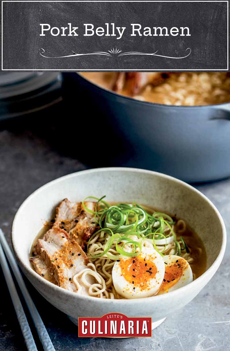 A white bowl filled with pork belly ramen, two egg halves, and scallions with a Dutch oven in the background and a pair of chopsticks lying beside the bowl.