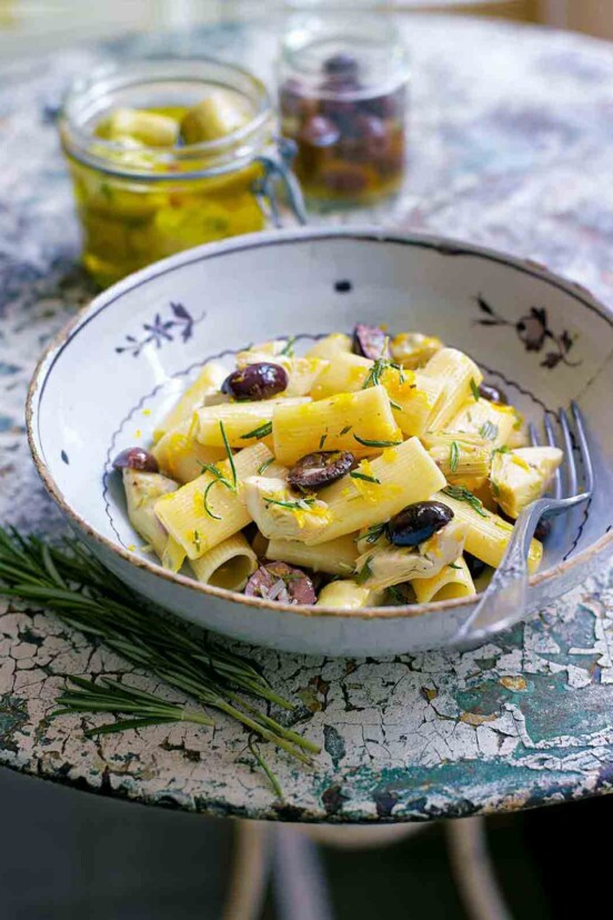 Bowl of rigatoni with artichokes, garlic, and olives, with lemon zest on a textured table