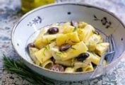 Bowl of rigatoni with artichokes, garlic, and olives, with lemon zest on a textured table