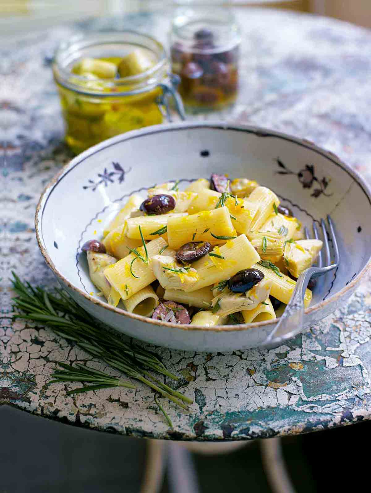 Bowl of rigatoni with artichokes, garlic, and olives, with lemon zest on a textured table