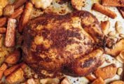 A whole roast chicken rubbed with paprika on a baking sheet with carrots and sweet potatoes.