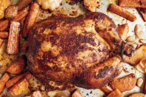 A whole roast chicken rubbed with paprika on a baking sheet with carrots and sweet potatoes.